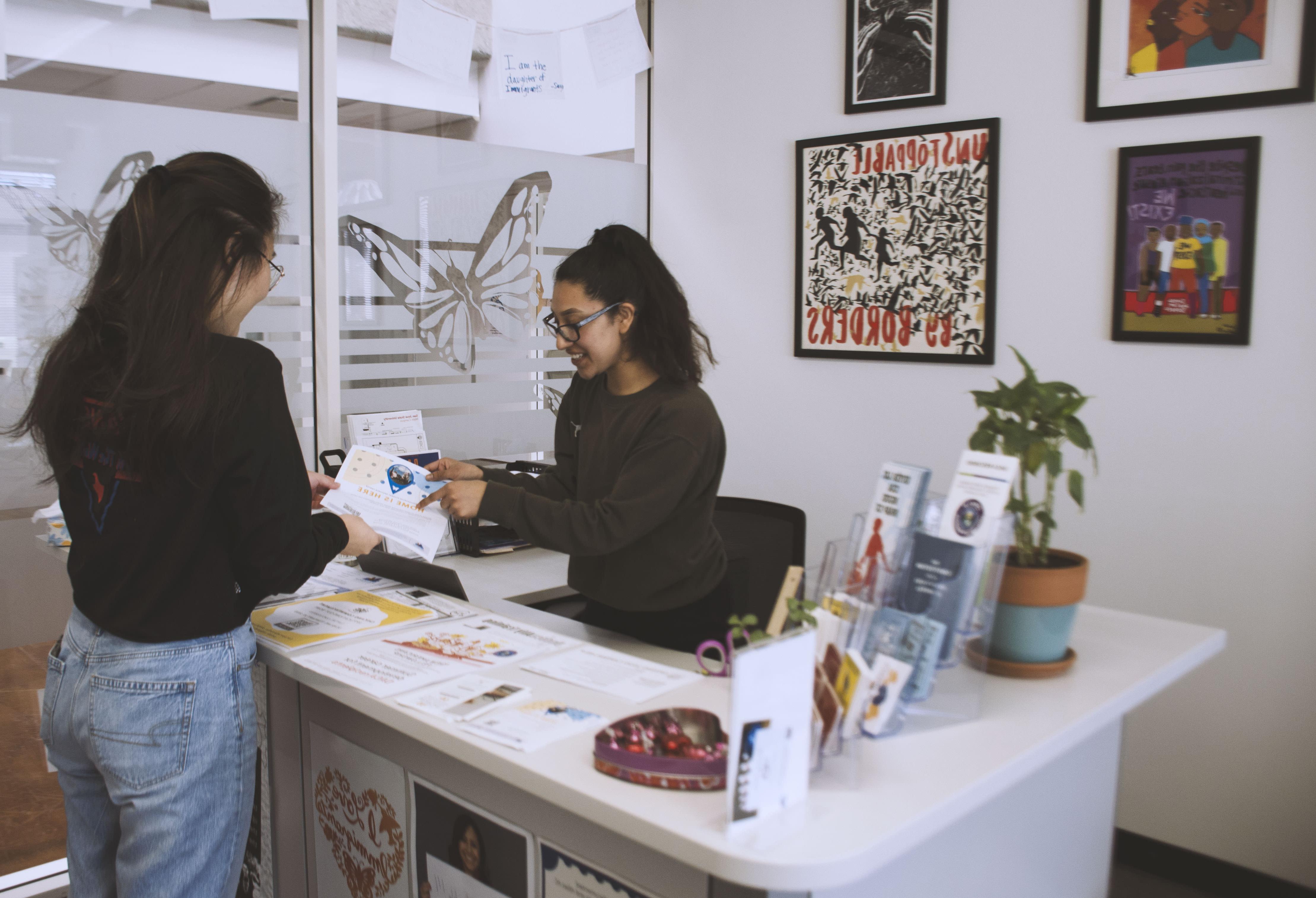 Front desk area of the UndocuSpartan Student Resource Center at San Jose State University, featuring two young students at the front desk with informational materials, a welcoming atmosphere, and a backdrop with decorations related to the center's mission.